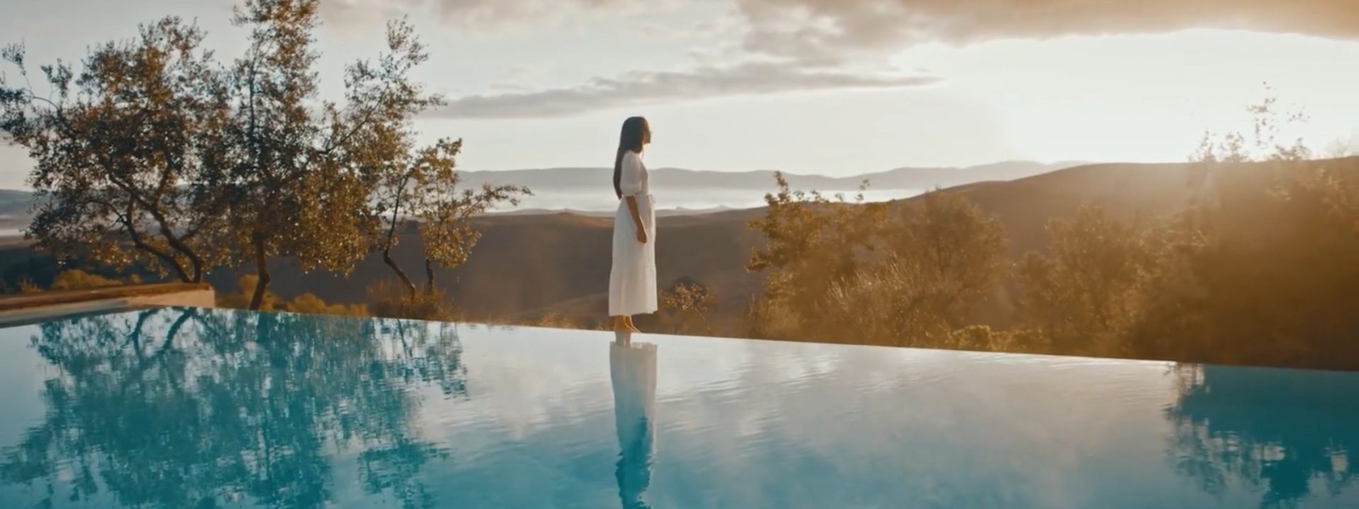 A woman in a white dress stands by an infinity pool, reflecting the serene landscape of trees and hills at sunset, creating a calm, peaceful scene.
