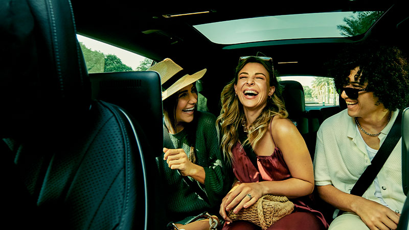Three young adults sit in the backseat of a car, smiling and laughing together during the daytime.
